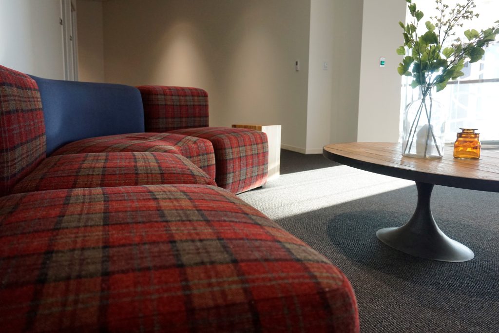 A room with a red and gray plaid-patterned sectional sofa on one side. A round wooden coffee table with a vase holding green flowers and a small amber jar is positioned in front of the sofa.