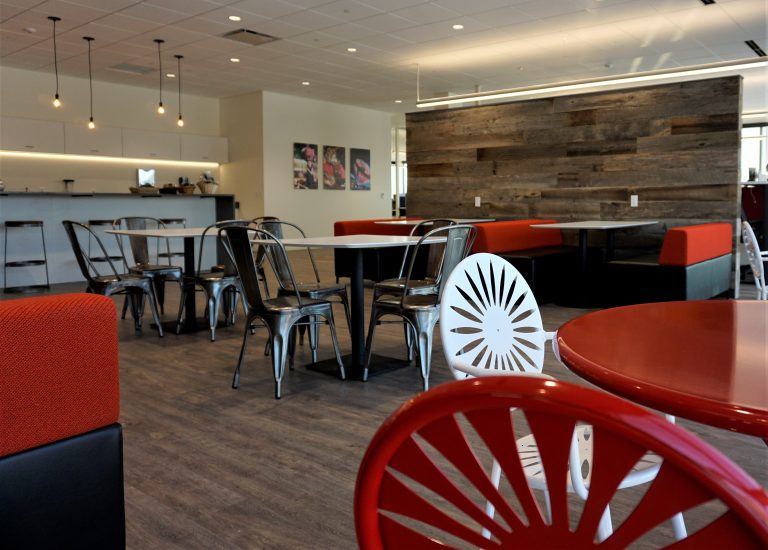 A modern cafeteria with a mix of red and white chairs, metal chairs, and red cushioned booth seating. The space features wooden accents, pendant lighting, and an open seating arrangement with a counter and kitchen area in the background.