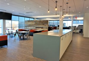 office kitchen and lounge area featuring a large countertop, hanging lights, and a mix of seating options including tables, chairs, and cushioned bench seats. Floor-to-ceiling windows provide ample natural light, and office cubicles are visible in the background.