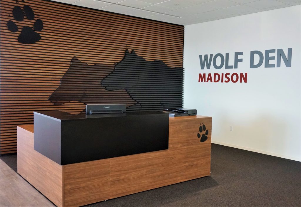 A modern office reception area featuring a wooden desk with a black countertop. Behind the desk is a wall with horizontal wooden slats and a wolf silhouette design.