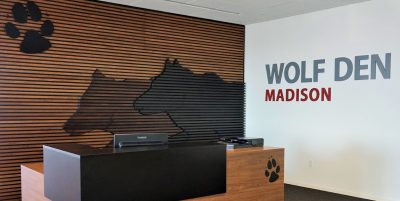 A modern office reception area featuring a wooden desk with a black countertop. Behind the desk is a wall with horizontal wooden slats and a wolf silhouette design.