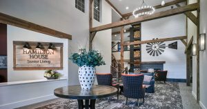 Senior Living facility lobby with gray patterned carpet, wood sign, and large fireplace with white horizontal shiplap