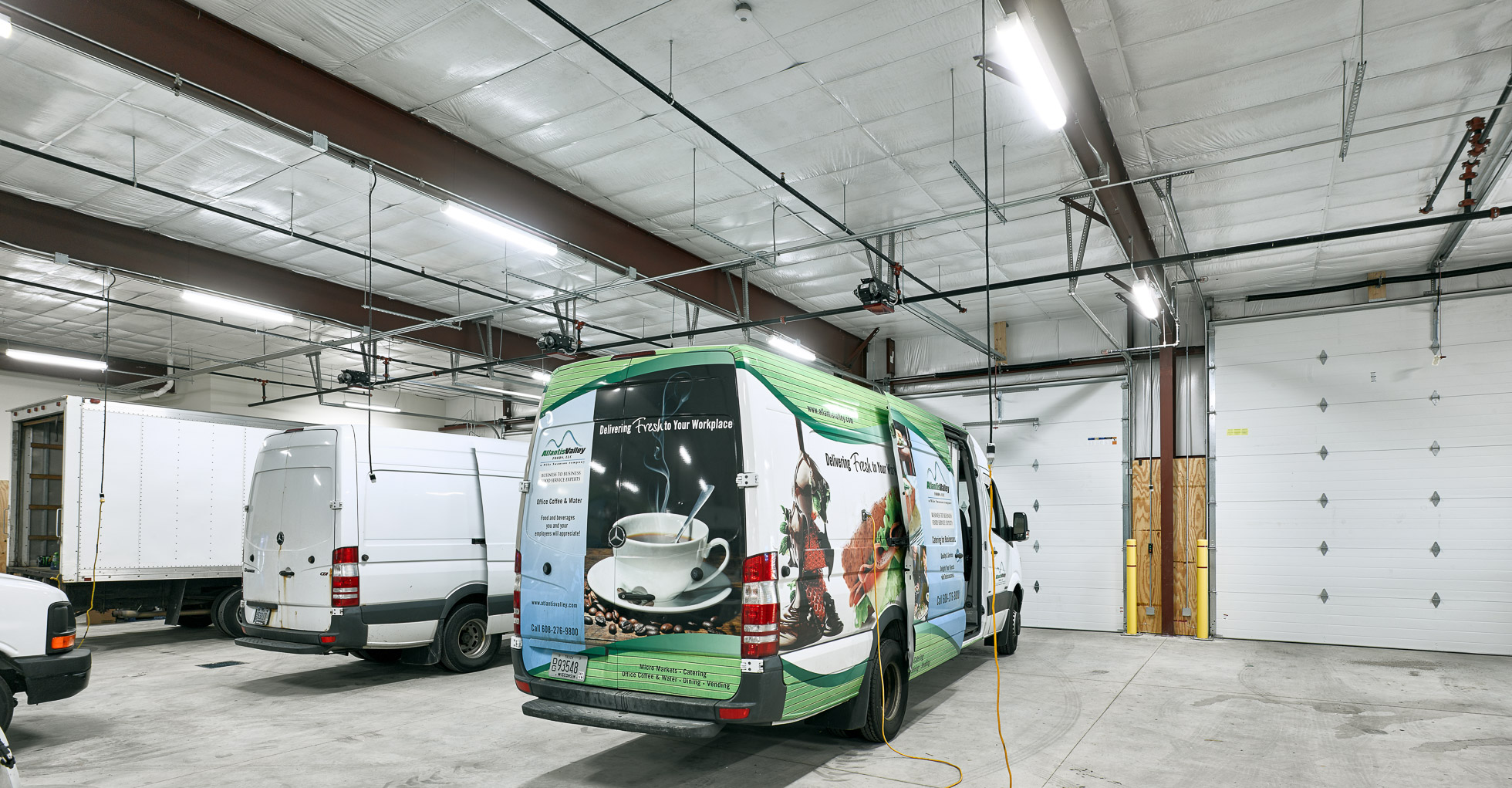 A spacious garage with three vans parked inside. One van features a colorful advertisement for coffee, displaying a cup and beans. The garage is well-lit with a high ceiling and several overhead doors.