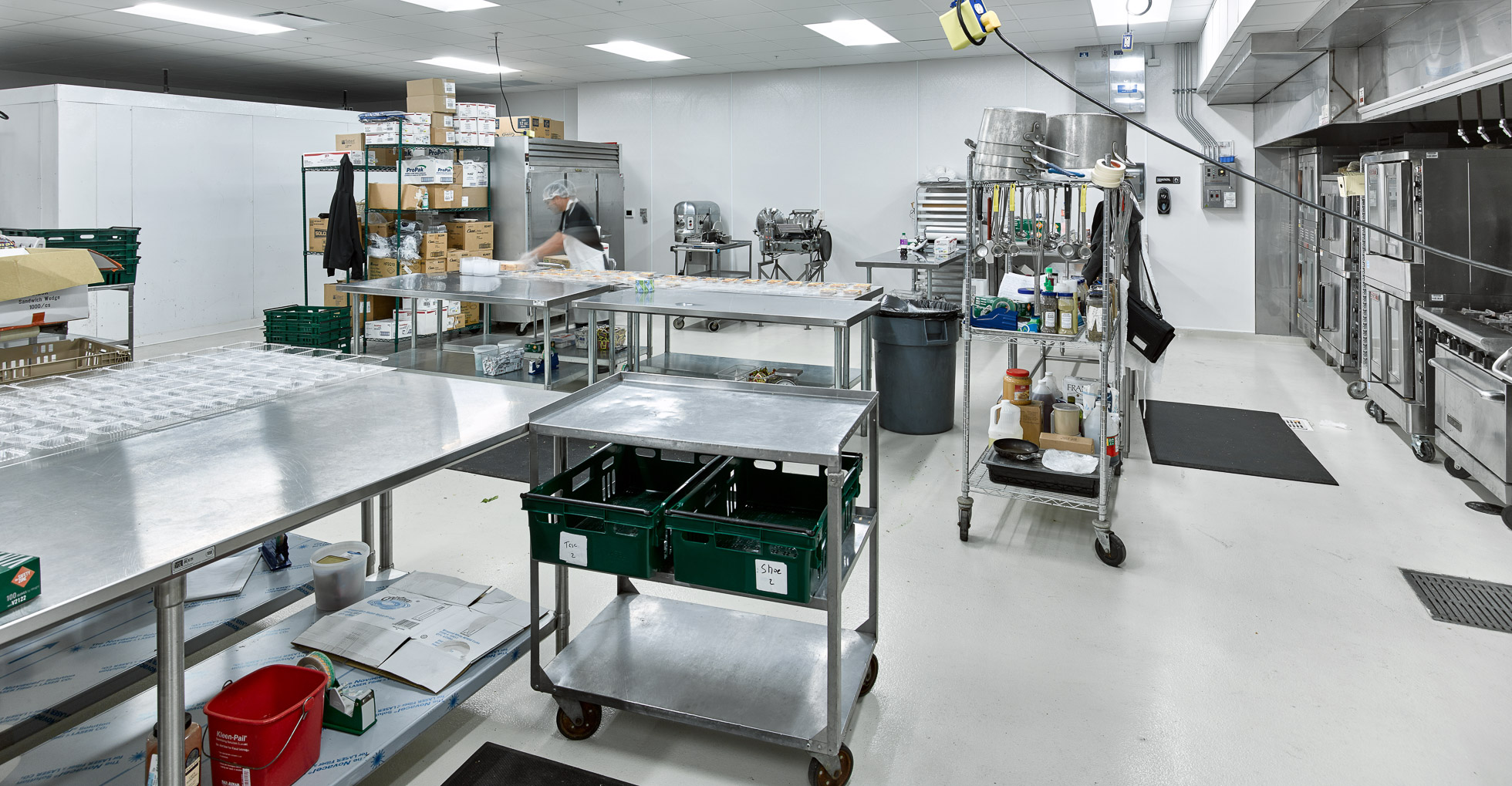 A commercial kitchen with stainless steel tables, shelves stocked with supplies, and industrial appliances. A person in the background is preparing food near trays and utensils. The area is clean and brightly lit.