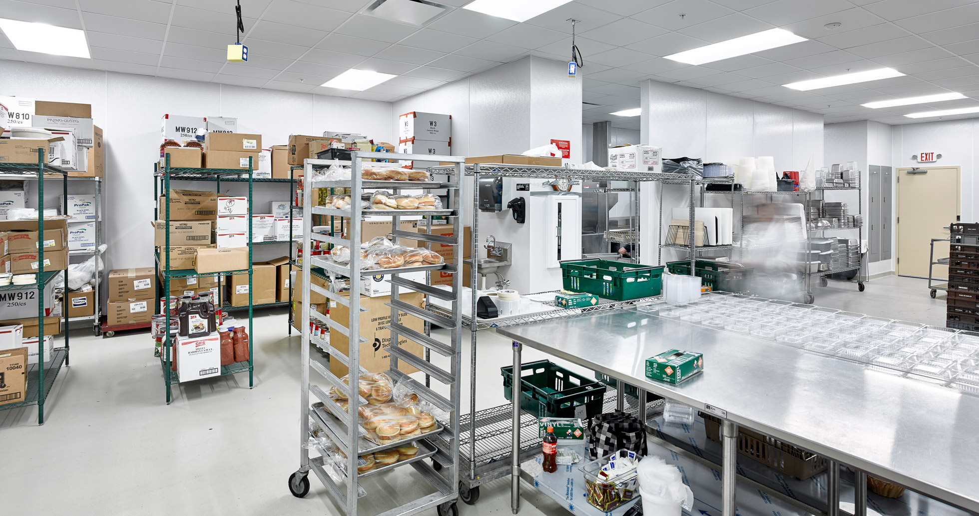 Industrial kitchen storage area with metal shelves and racks holding boxes, trays of pastries, and various supplies. Stainless steel tables are in the center, with a blurred motion figure suggesting movement. Bright overhead lighting illuminates the room.