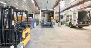 A warehouse interior with a forklift, a delivery truck being loaded, and a green van. The space is filled with pallets of bottled goods, arranged neatly along the sides. Overhead fluorescent lights illuminate the large open area.