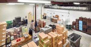 A warehouse interior filled with stacked boxes, packaged goods, and beverages on shelves. A person in the back is operating a hand truck. Ceiling ductwork and an overhead light are visible, creating an organized storage space.