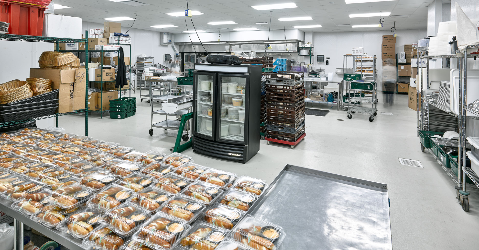 A commercial kitchen with stainless steel tables and equipment. Packaged food items are neatly arranged on a table in the foreground. Shelving units hold various supplies, and a refrigerator stands to the center. The area is well-lit and organized.