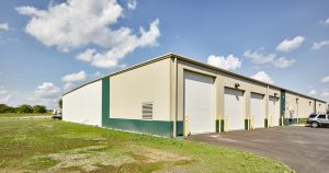 A large beige industrial building with several garage doors sits under a blue sky with scattered clouds. The building is surrounded by a grassy area and asphalt driveway, with a few vehicles parked near the entrance.