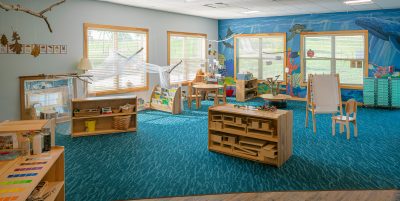 A spacious classroom with vibrant blue carpet featuring various learning stations and educational materials. Wooden furniture is scattered around, including shelves with toys and supplies, a small table with chairs, and an easel. Colorful decorations and artwork adorn the walls.