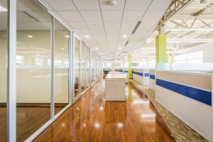 office collaboration area with long standing table with natural wood flooring glass walls and gray with a blue strip cubicle