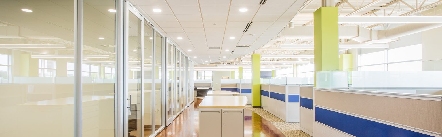 office collaboration area with long standing table with natural wood flooring glass walls and gray with a blue strip cubicle