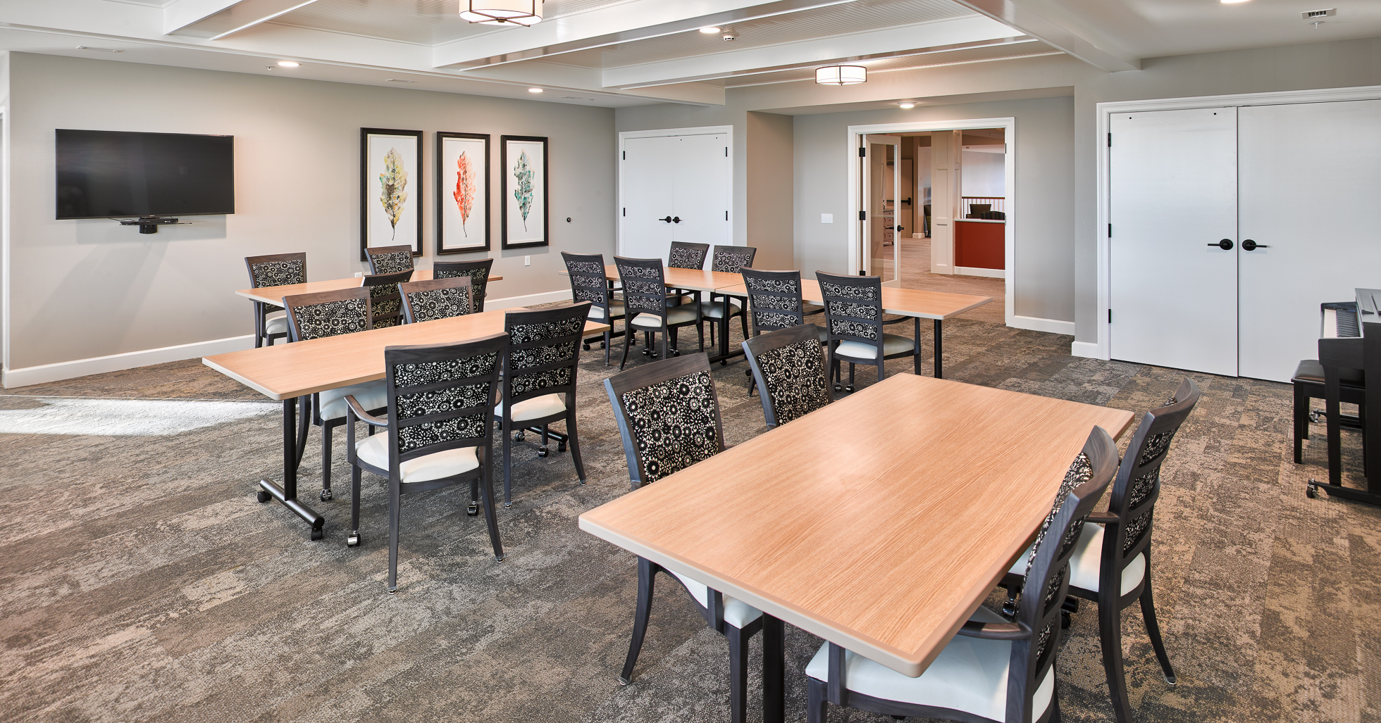 activity room with wood top tables and chairs and tv mounted on cream colored walls and a piano in the right corner