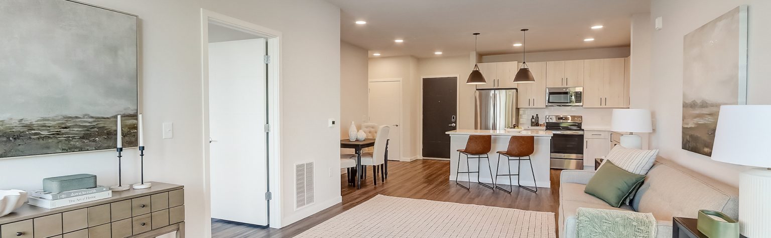 Apartment with light wooden cabinets, stainless steel appliances, an island with a white countertop, and two pendant lights hanging above. The kitchen opens to a living area with light-toned furniture.