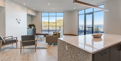 A modern, open-concept apartment community room with large windows offering a scenic view of a lake, natural wood flooring, and a kitchen island with a stone countertop