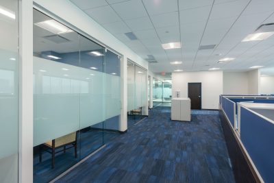 A modern office area with blue carpet and white walls. The space includes glass-walled meeting rooms on one side, and open cubicles with blue dividers on the other. Fluorescent lights illuminate the area, and an emergency exit sign is visible in the background.