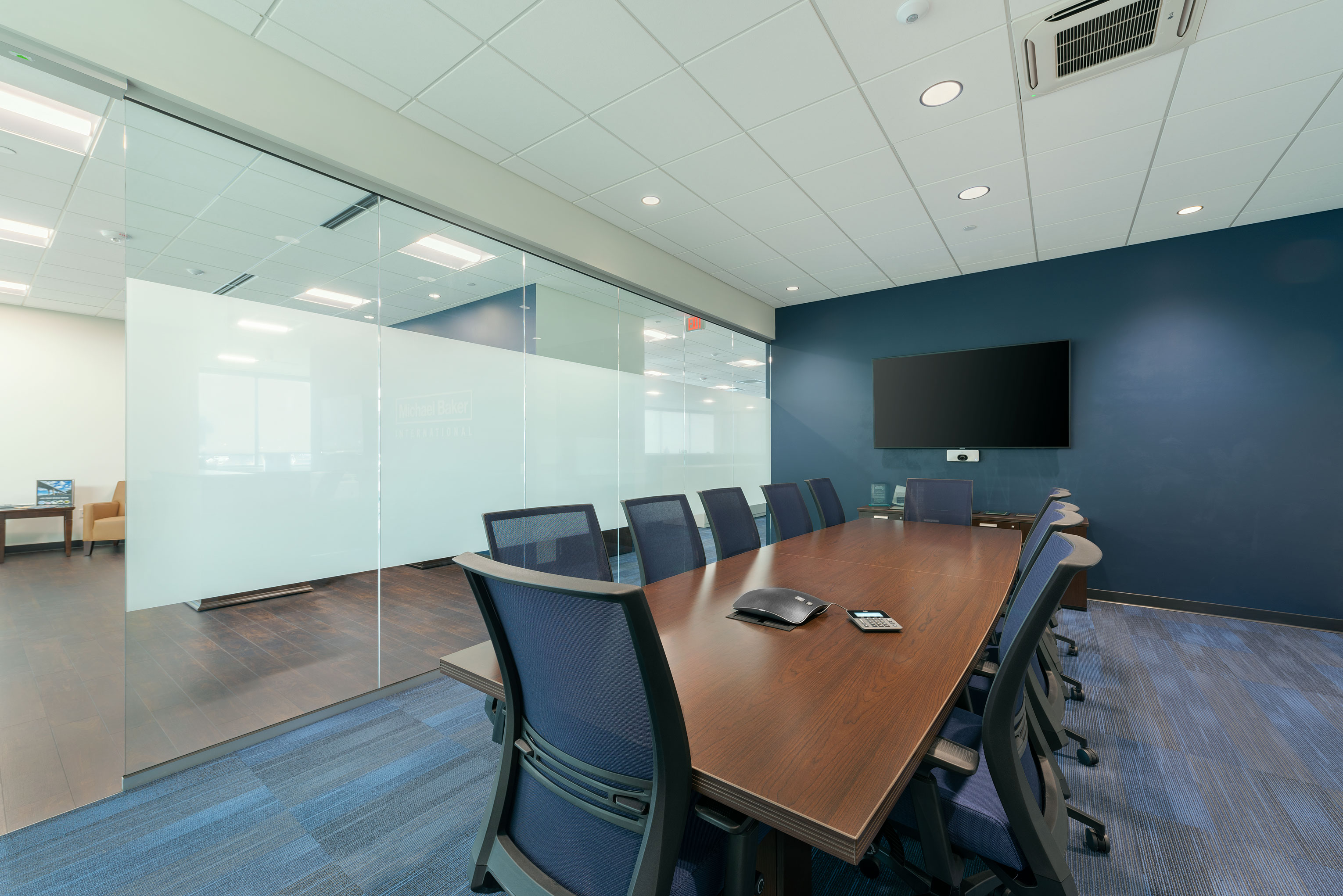 A modern conference room features a long wooden table surrounded by blue office chairs. A large flat-screen TV is mounted on a dark blue accent wall. The room has glass walls, allowing a view of a bright, open common area with light-colored furniture.