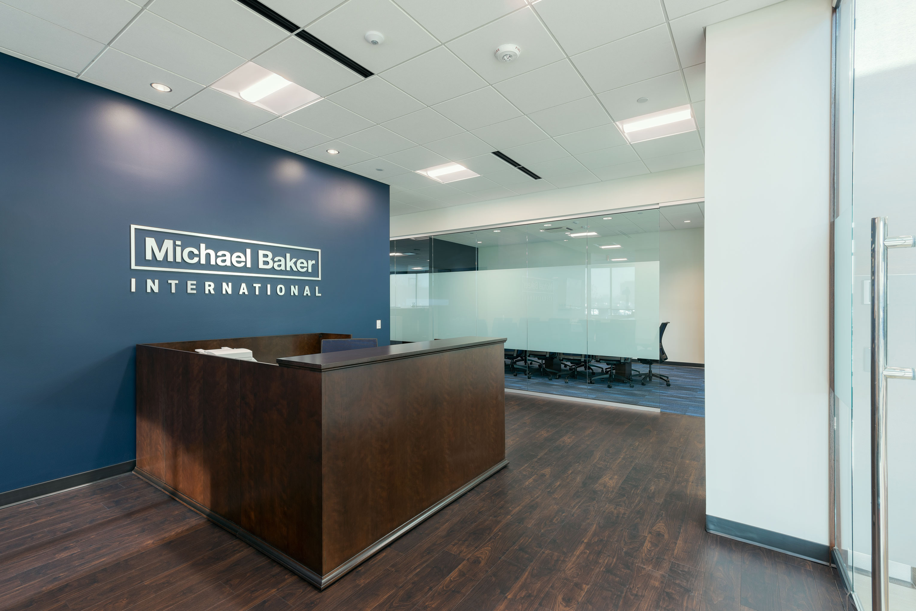 Modern office reception area with a dark wood desk in front of a blue wall displaying the text "Michael Baker International." The space features a glass wall partition and a meeting room in the background with several black office chairs around a table.