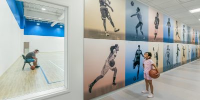 A young girl holding a basketball stands in a hallway, gazing at a wall adorned with large photos of athletes. Through a window to her left, a man is seated on a chair inside a basketball court, holding a basketball.