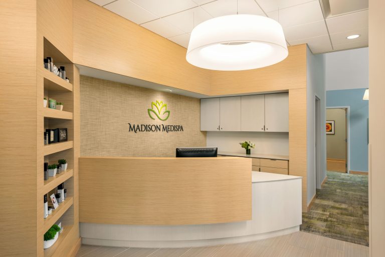 A modern, well-lit medical spa reception area with light wood cabinetry and a curved front desk. Behind the desk, the wall showcases the "Madison Medispa" logo with a leaf design. Shelves with products are on the left, and a hallway extends to the right.