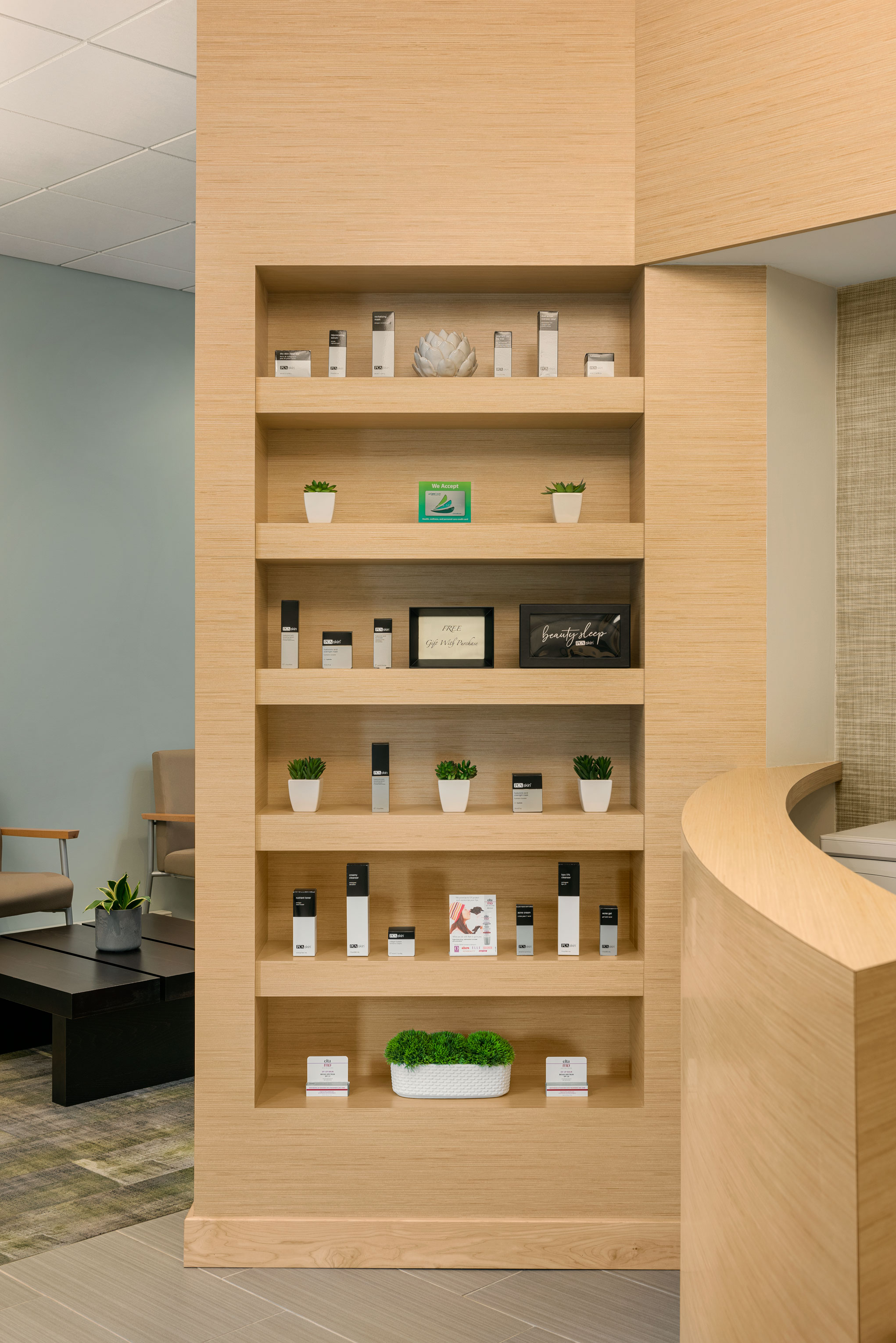 A light-colored wooden shelf built into a wall holds various skincare products and small potted plants. The shelf is neatly organized and displayed against a softly lit, modern interior space. At a Spa.