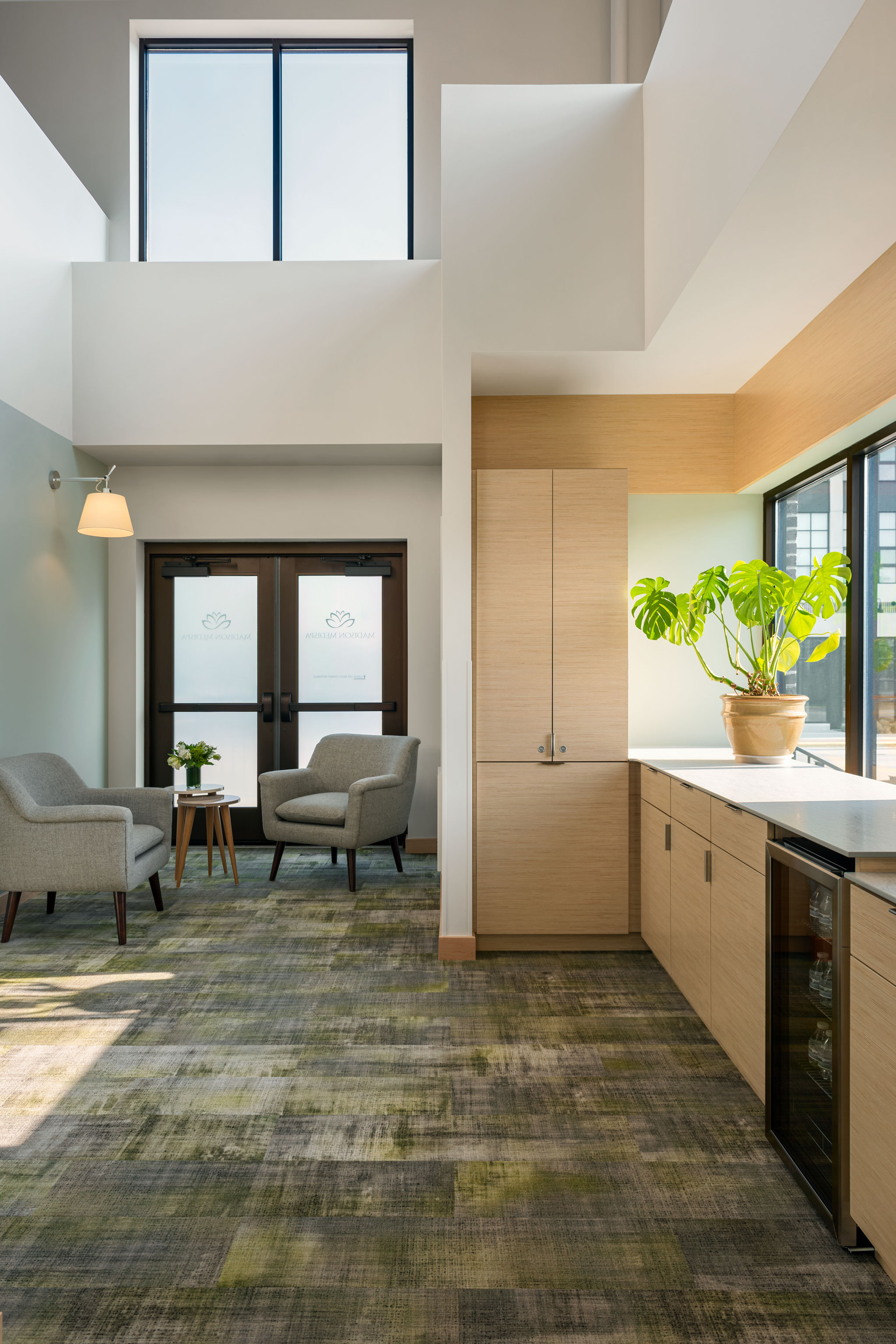 A modern, well-lit waiting area with two grey armchairs and a small wooden table topped with a plant and magazines. Adjacent is a kitchenette with light wooden cabinetry, a countertop holding a large plant, and a wine cooler. Large windows offer natural light.