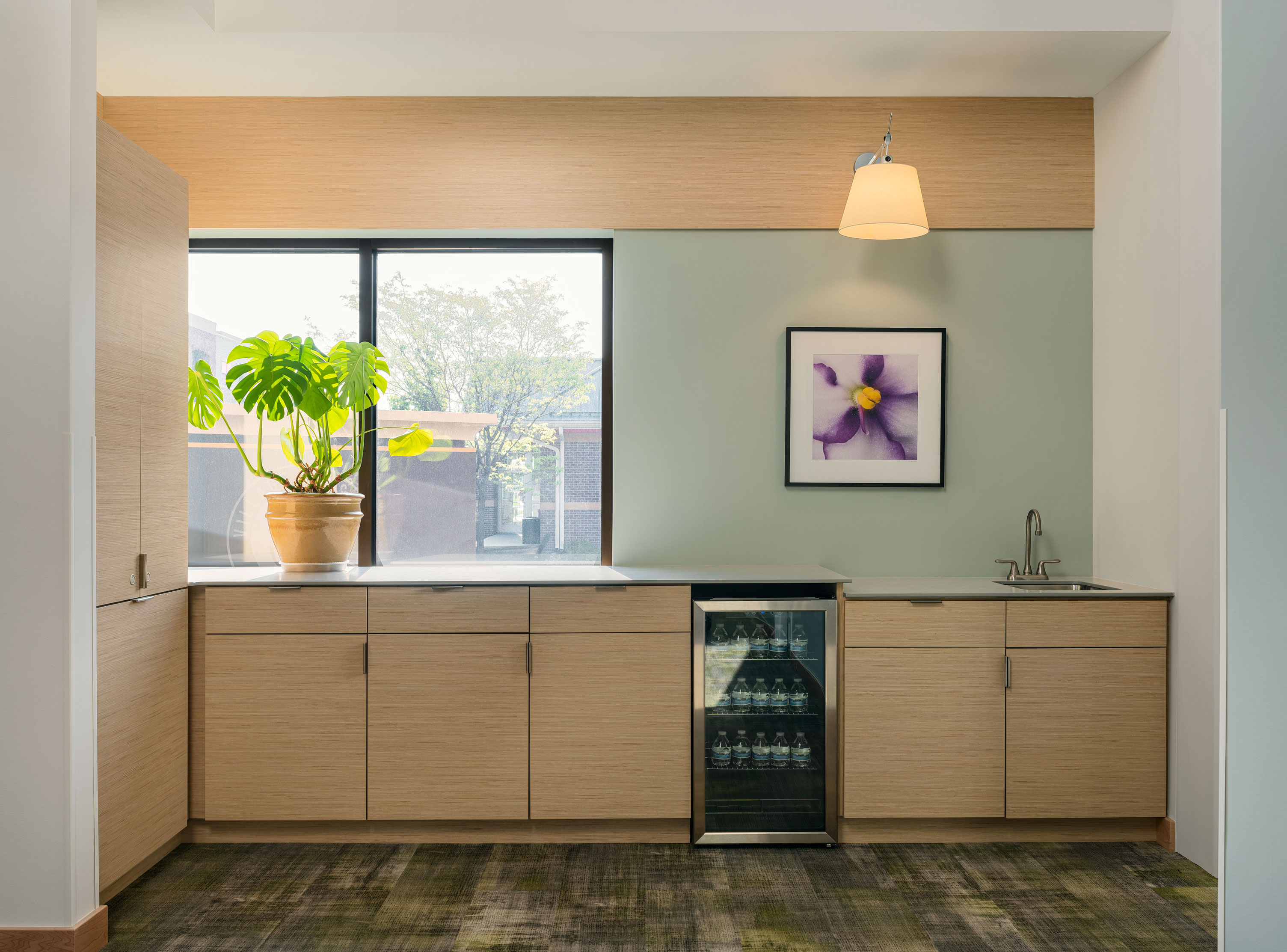 A modern kitchenette with light wood cabinetry, a small fridge with glass doors, a sink, and a potted plant by the window. Above the counter, there's a framed picture of a purple flower and a hanging light fixture. The floor features a patterned carpet.