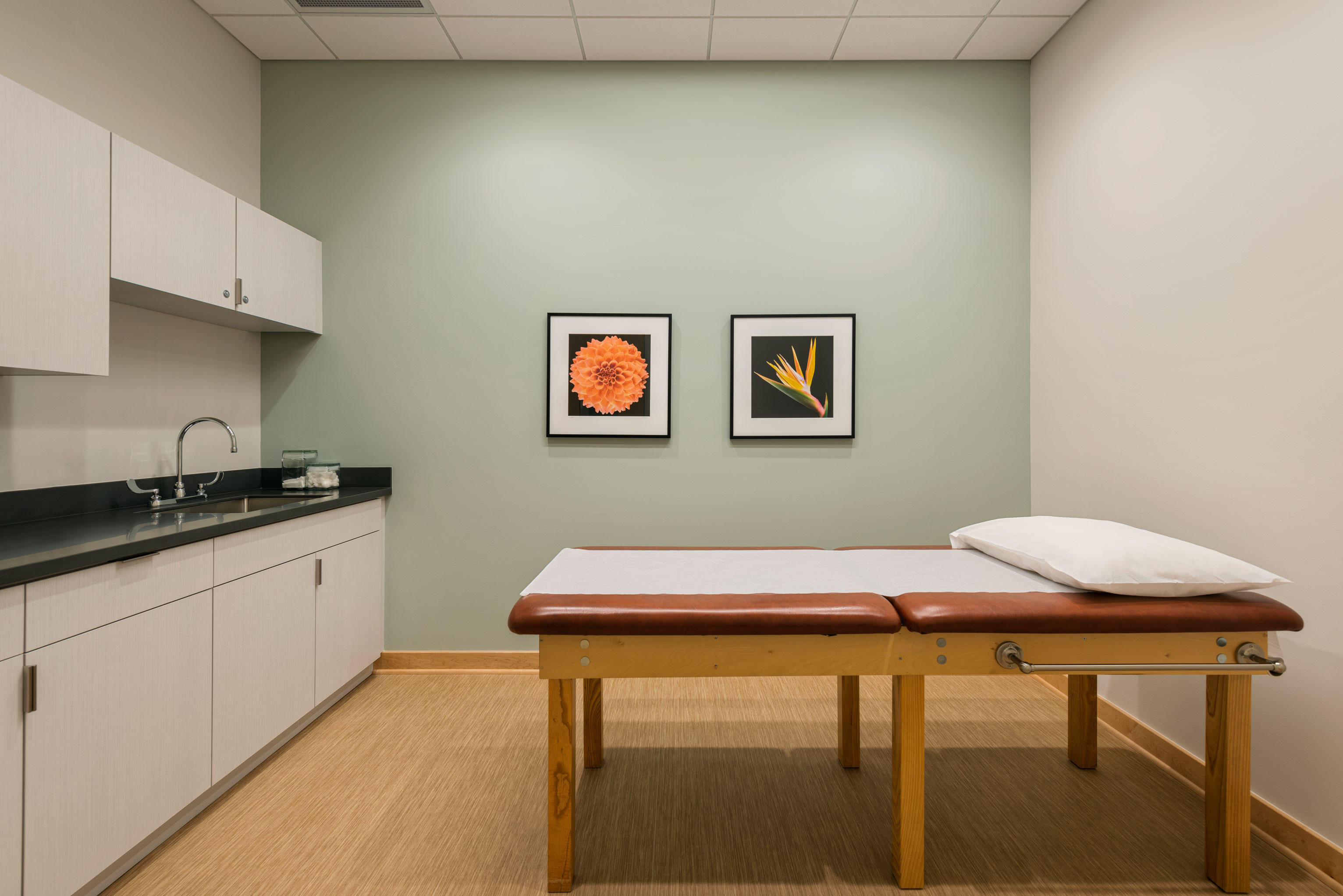 A clean and well-lit examination room with a wooden treatment table topped with a white pillow in the center. The walls are light green and white, adorned with two framed floral pictures. A counter with cabinets and a sink is on the left side of the room.