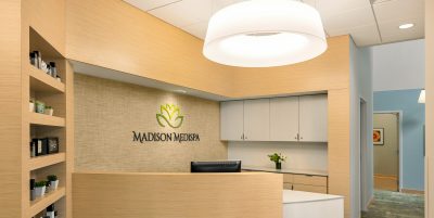 A modern, well-lit medical spa reception area with light wood cabinetry and a curved front desk. Behind the desk, the wall showcases the "Madison Medispa" logo with a leaf design. Shelves with products are on the left, and a hallway extends to the right.