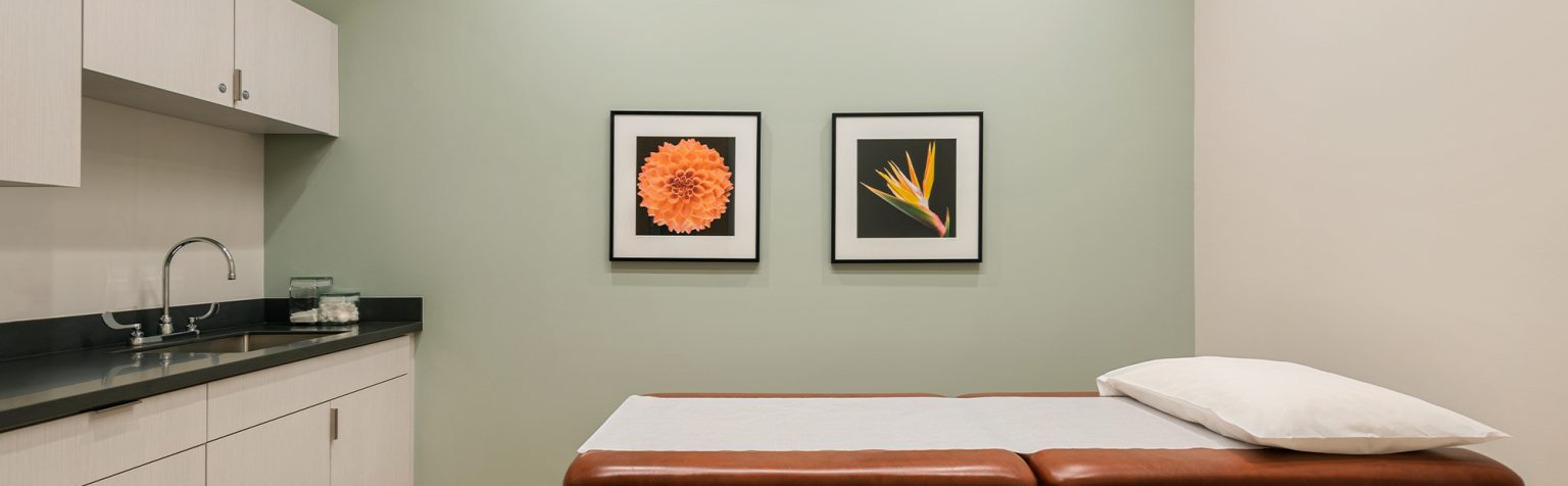 A clean and well-lit examination room with a wooden treatment table topped with a white pillow in the center. The walls are light green and white, adorned with two framed floral pictures. A counter with cabinets and a sink is on the left side of the room.