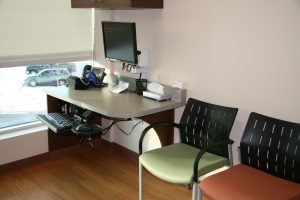 A doctor's office features a wall-mounted computer with keyboard and mouse on a desk. Below the desk is a blood pressure cuff. Two chairs, one green and one black, are positioned beside the desk. A large window lets in natural light, revealing cars parked outside.