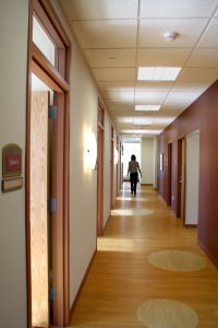 A person walks down a brightly lit corridor with wooden floors and beige and brown walls. Several doors line the hallway, each with a nameplate, including one labeled "Exam 2". Circular light fixtures are mounted on the walls, and natural light comes in from a window at the end.