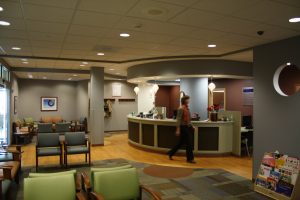 A modern waiting room with comfortable chairs, a circular reception desk, and a person walking behind it. The room is well-lit with natural light from large windows and has a magazine stand on the right. Coats hang on wall hooks near the reception area.