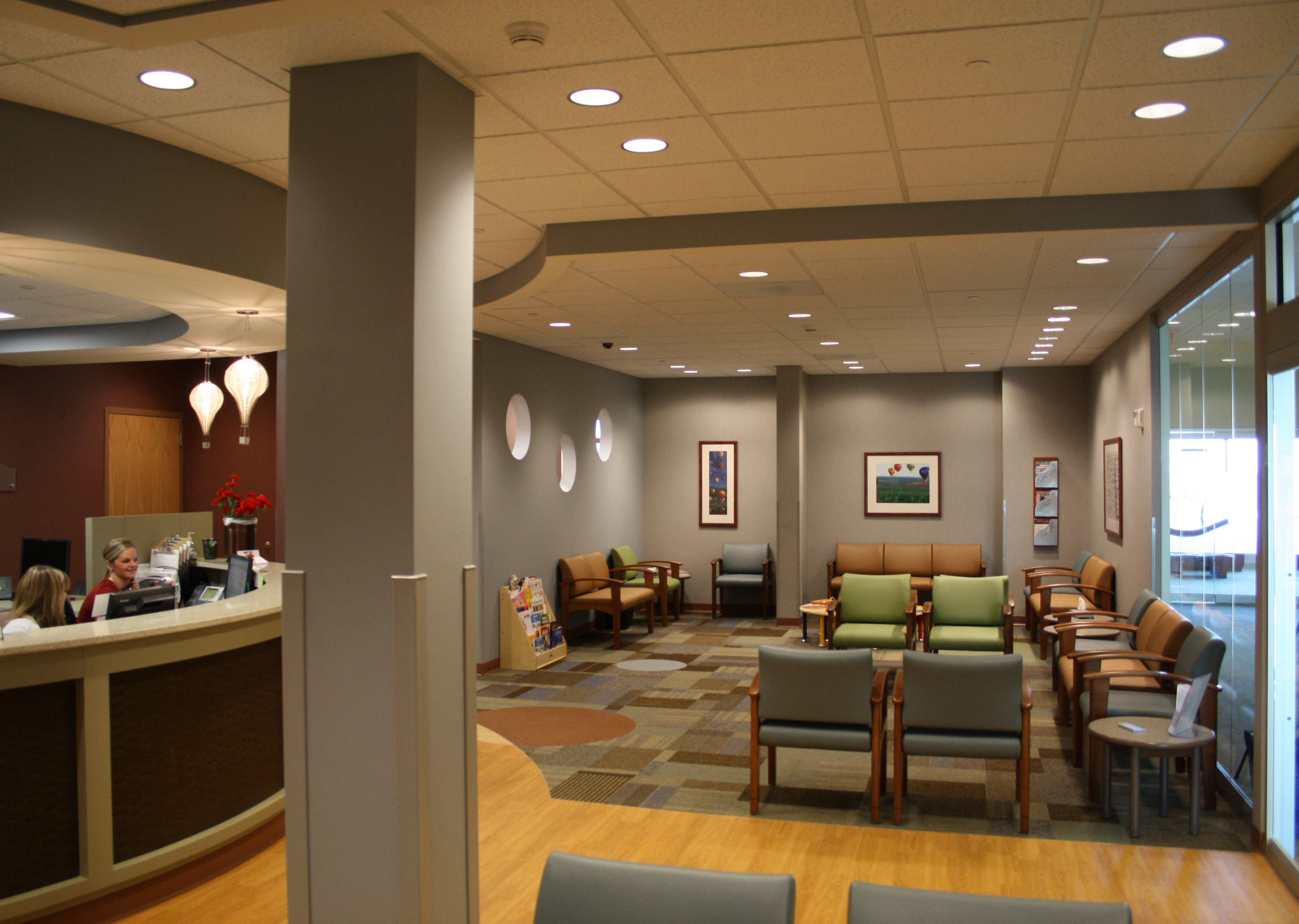 A modern, well-lit waiting room with comfortable seating, artwork on the walls, and a reception desk on the left with staff members attending to visitors. The room has a light-colored ceiling and large, clear windows allowing natural light to enter.