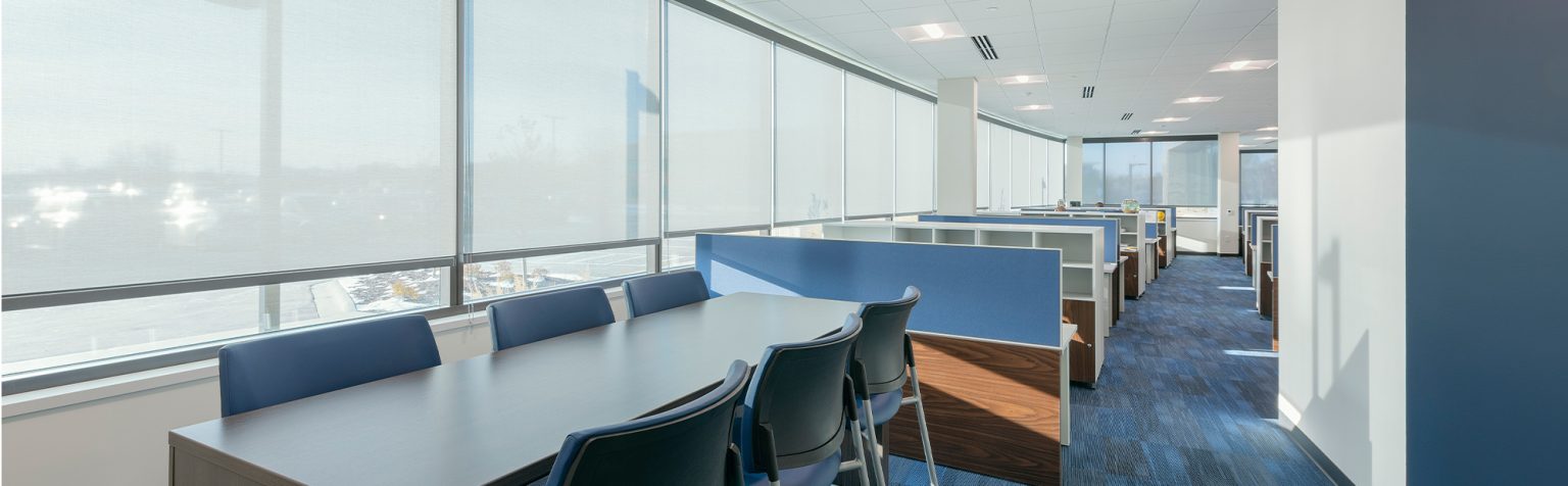 office space with blue carpet and white ceilings features long tables with chairs for collaborative work, and individual cubicles along the side. Large windows allow natural light to flood in, offering a bright and open atmosphere.