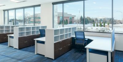 office space with large windows allowing natural light to illuminate the room. The office features rows of desks with blue chairs and white shelving. Outside the windows, a landscape of trees and buildings can be seen. The flooring is blue carpet.