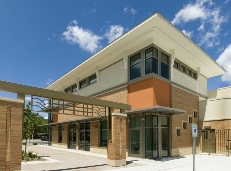 A modern two-story building with a mix of beige and orange walls, large windows, and a decorative metal awning. The sky is clear with a few clouds, and there’s a handicap parking sign in the foreground.