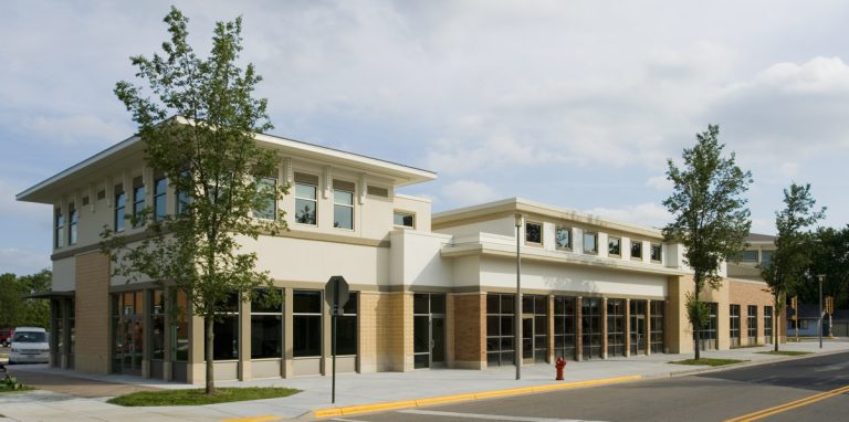 A modern two-story commercial building with large windows and beige exterior is situated on a corner. It is surrounded by young trees and a paved street. The sky is partly cloudy.