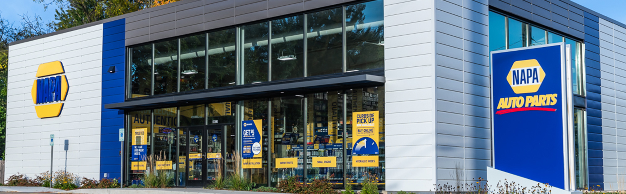 A modern building with a blue and gray facade featuring the NAPA Auto Parts logo. The front has large glass windows and doors with promotional signs.