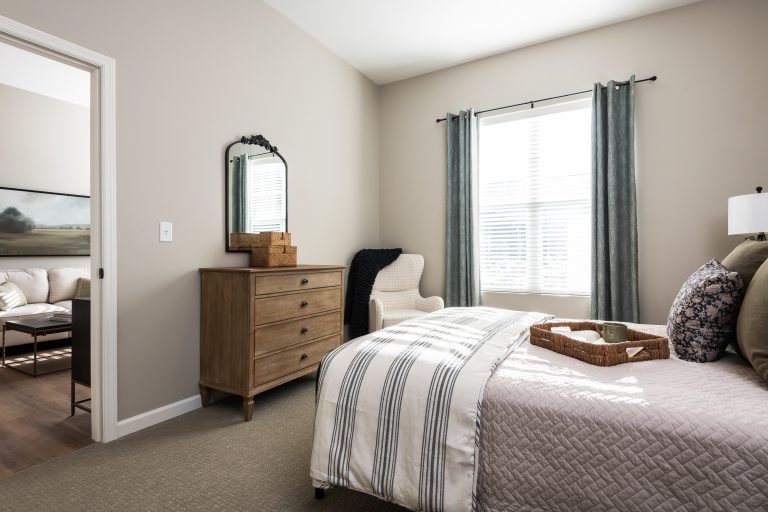 Assisted living bedroom with a bed covered in a striped blanket and decorative pillows. A wooden dresser with a mirror and a woven basket sits beside a white armchair.