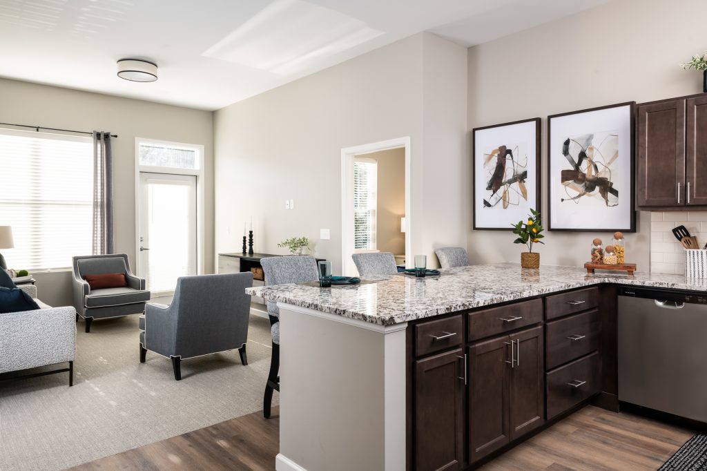 Modern kitchen and living area with granite countertops and dark wood cabinets. The living area features gray chairs and a large window with natural light. Abstract art hangs on the wall above the counter.