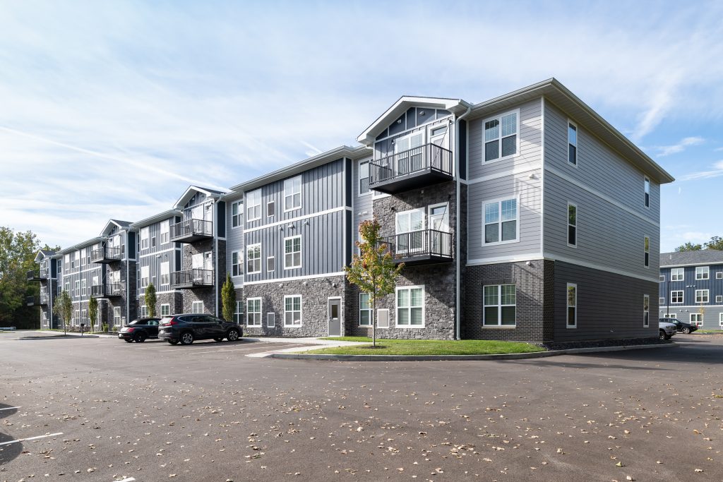 Exterior of assisted living apartments with gray siding and balconies, set against a blue sky. Scattered leaves on the paved parking area; a few cars are parked. Sparse trees and grass surround the buildings.