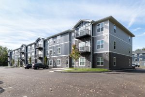 Exterior of assisted living apartments with gray siding and balconies, set against a blue sky. Scattered leaves on the paved parking area; a few cars are parked. Sparse trees and grass surround the buildings.