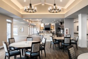 spacious dining area with several round tables and chairs, wooden flooring, and large chandeliers hanging from the ceiling. The room is well-lit with natural light from large windows