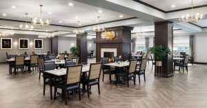 Community room with wooden floors and black-and-beige chairs around rectangular tables. There's a fireplace in the center, surrounded by dark wood panels. Elegant chandeliers hang from the ceiling, and framed art decorates the walls.