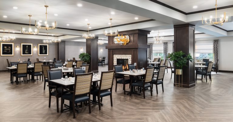 Community room with wooden floors and black-and-beige chairs around rectangular tables. There's a fireplace in the center, surrounded by dark wood panels. Elegant chandeliers hang from the ceiling, and framed art decorates the walls.