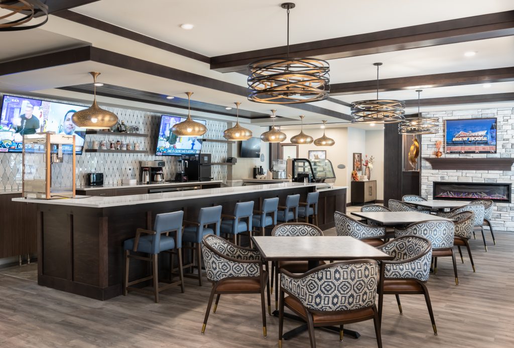 Dinning room and lounge with a bar, high stools, and several tables. Multiple TV screens are mounted on the wall above the bar. Pendant lights hang from the ceiling, and a stone fireplace is in the corner.