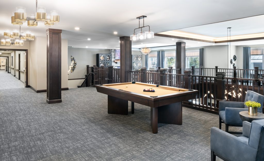 A modern interior featuring a billiards table with balls arranged for a game. The space has elegant lighting, dark wood accents, and light gray carpet. Two gray chairs with a small table and potted plant are nearby. Large windows bring in natural light.