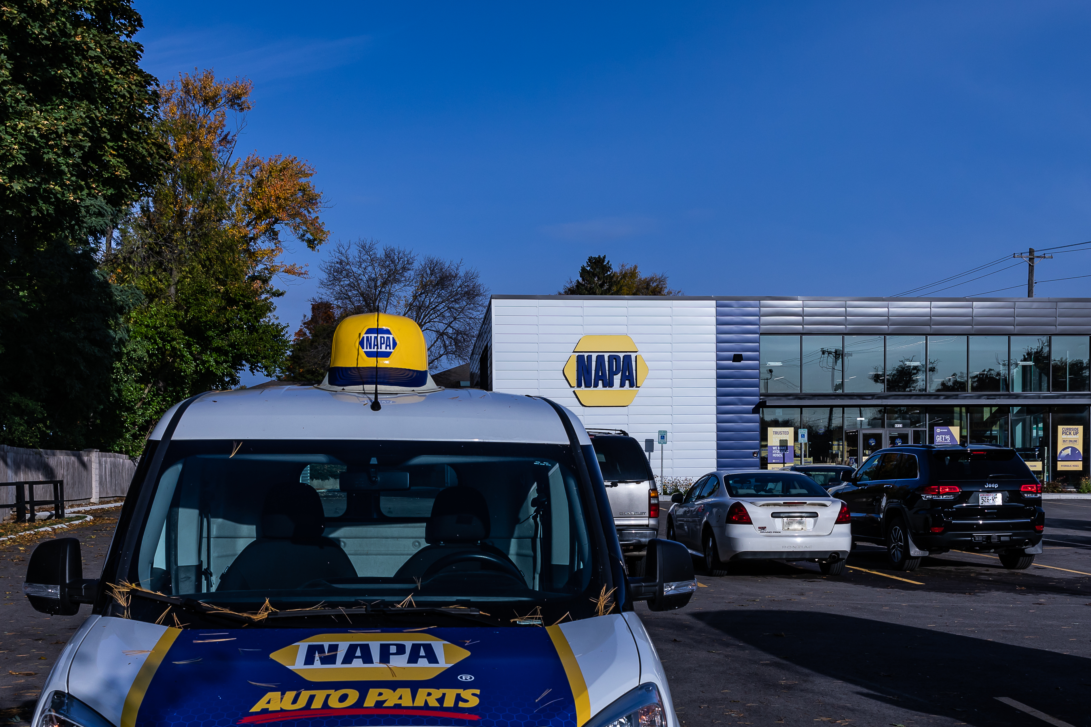 A parking lot with several cars is in front of a NAPA Auto Parts store. A NAPA Auto Parts van is parked in the foreground, displaying the store's branding. The sky is clear, and trees with autumn leaves are visible.