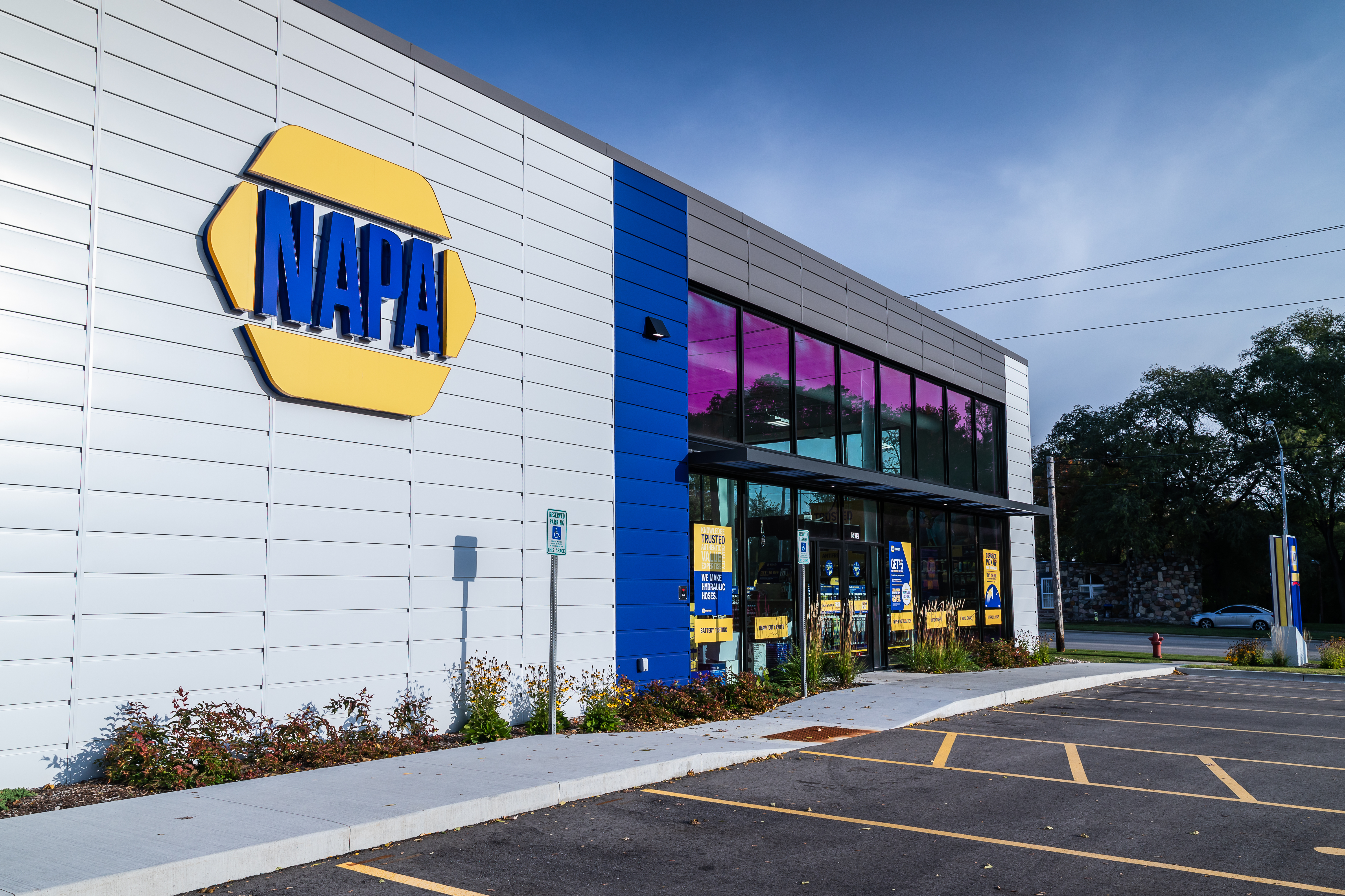 Side view of a NAPA Auto Parts store with a modern exterior featuring a large logo. The building has blue and grey panels with glass windows. A parking area with marked spaces is visible in the foreground. Trees line the background under a partly cloudy sky.